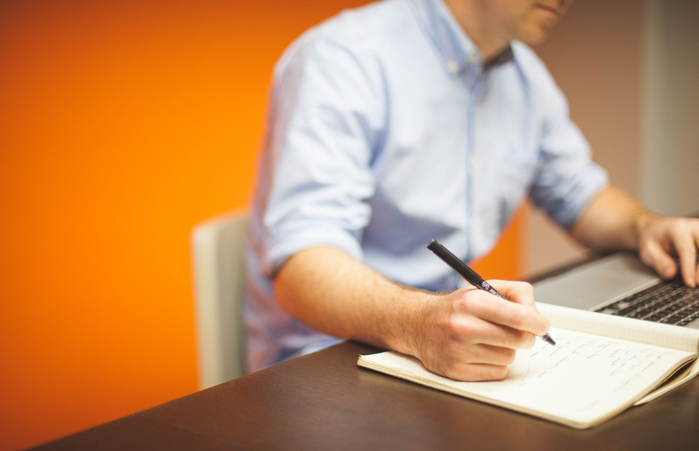 businessman-man-space-desk-7059 orange background; person in collared blue shirt; one hand on laptop and other writing in notepad