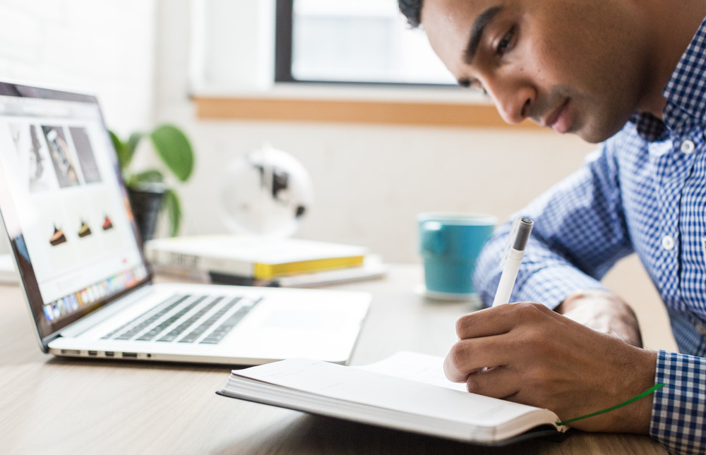 man-using-ballpoint-pen-374820 person in checkered blue shirt writing on notepad; laptop in front
