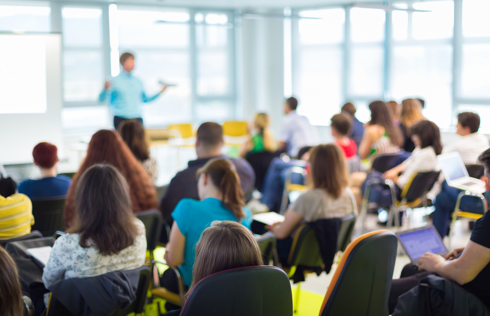 shutterstock_206744158 small meeting room with speaker in blue shirt, blurred; speaker speaking to seated audience taking notes