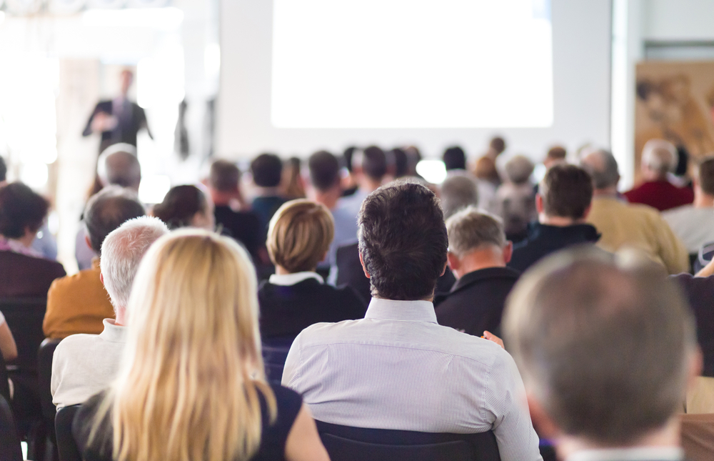 conference room with audience listening to speaker in front; audience seated, speaker standing