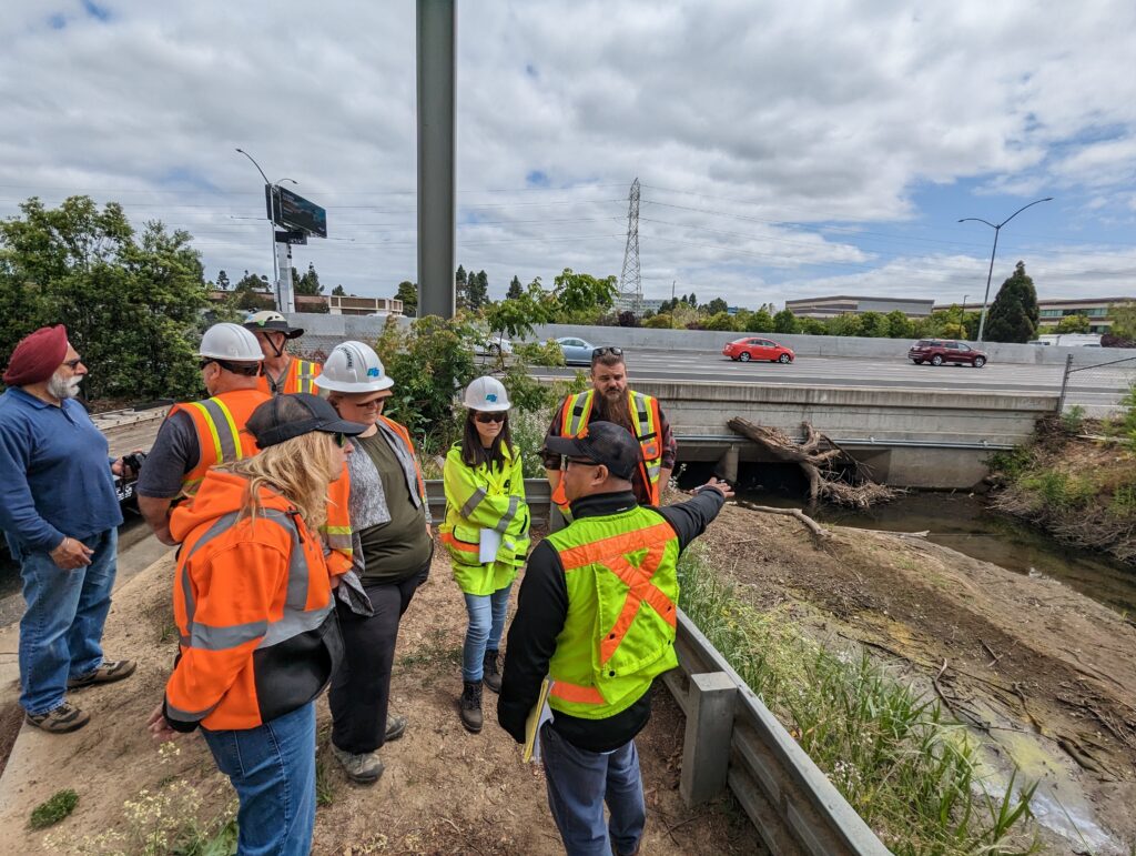 A group of people wearing safety vests and hard hats looking at a creek with debris
