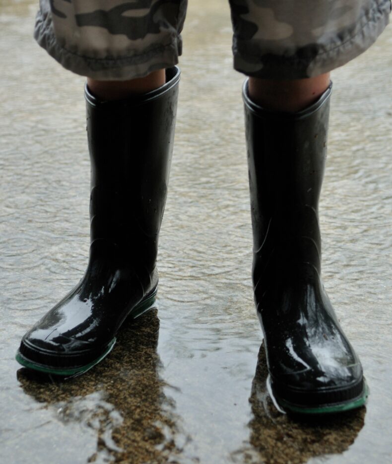 Close-up of a person standing in shallow water, wearing black rubber rain boots and rolled-up camouflage-pattern pants. The boots are partially submerged on a wet concrete or paved surface, suggesting flooding or heavy rain conditions.